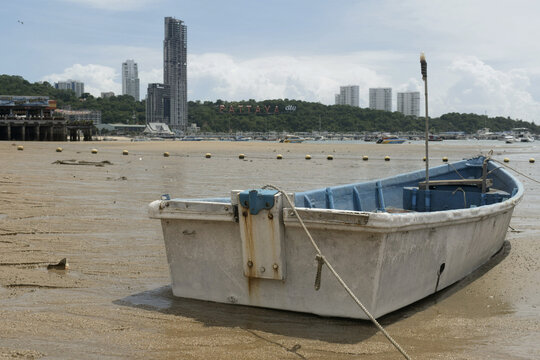 small boat kept on the sandy shore of pattaya beach during low tide with the city skyline and coastal buildings visible in the background
