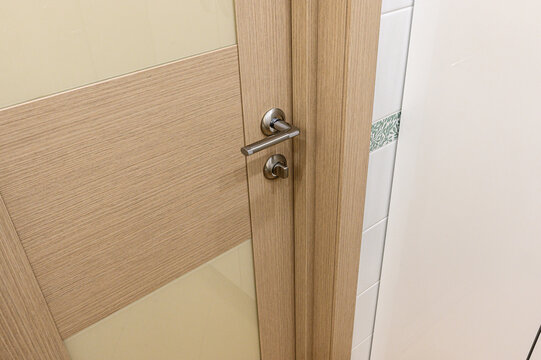 A light‑oak interior door features a textured wood‑grain panel and a sleek brushed‑metal lever handle with a matching lock. it frames a tiled wall with a subtle decorative strip