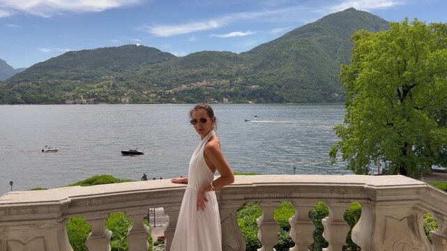 Beautiful Confident Woman in Sleeveless White Halter Dress and Sunglasses on a Baroque Stone Terrace of a Historic Italian Villa With a Stunning Panoramic View of Lake Como and Alpine Mountains, Lomba