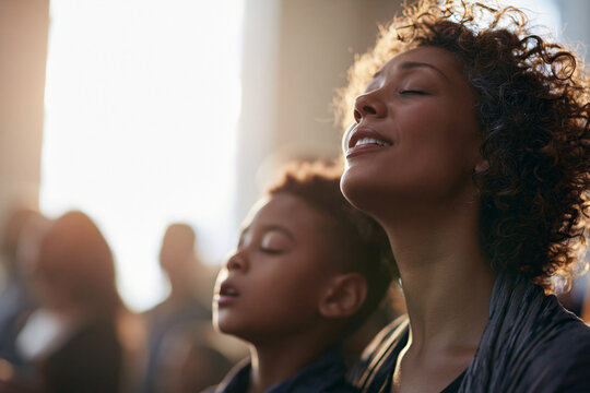 Devout African American mother and son with closed eyes singing and praying during a modern Christian church worship service, warm sunlight.
