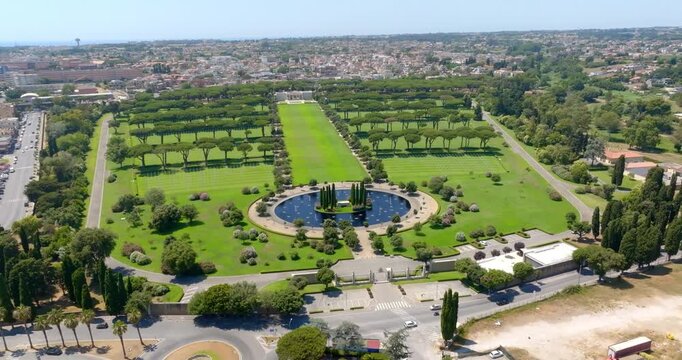 Wide aerial view of the Sicily Rome American Cemetery in Nettuno, showing a large circular pond with water lilies, green lawns, rows of pine trees, and the city in the background.