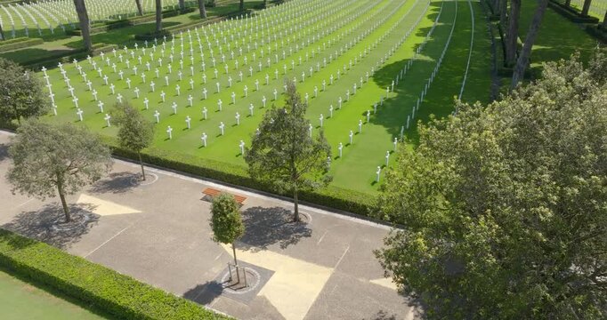 High angle view of a military cemetery with rows of white crosses on green grass, bordered by trees and a paved walkway. Aerial panorama of an American military cemetery in a sunny morning.