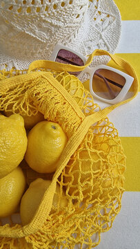  Summer vacation, summertime, travel, tourism concept. Top view of fresh lemons in a yellow mesh bag with a white sun hat and sunglasses on a yellow and white striped beach towel.