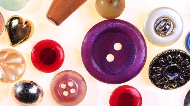 Assorted vintage buttons spread on cream paper background, showing pink, purple, red, gold and silver pieces in various shapes and finishes. Camera pans to right
