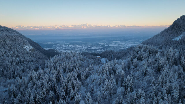 Aerial view of Col de la Faucille featuring snow-covered pine forests and distant mountain peaks under a soft sunset sky in Gex, Auvergne-Rhone-Alpes, France.