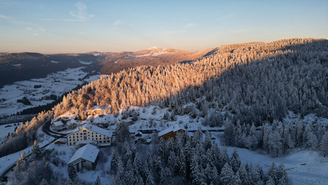 Aerial view of Col de la Faucille during winter, featuring snow-covered pine forests, mountain slopes, and alpine buildings under a clear sky in Gex, Auvergne-Rhone-Alpes, France.