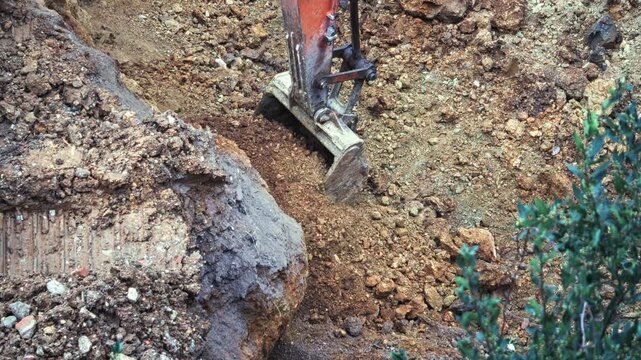 A backhoe excavator bucket digging into and scooping up rocky soil and dirt at a construction site foundation base. Close-up shot 