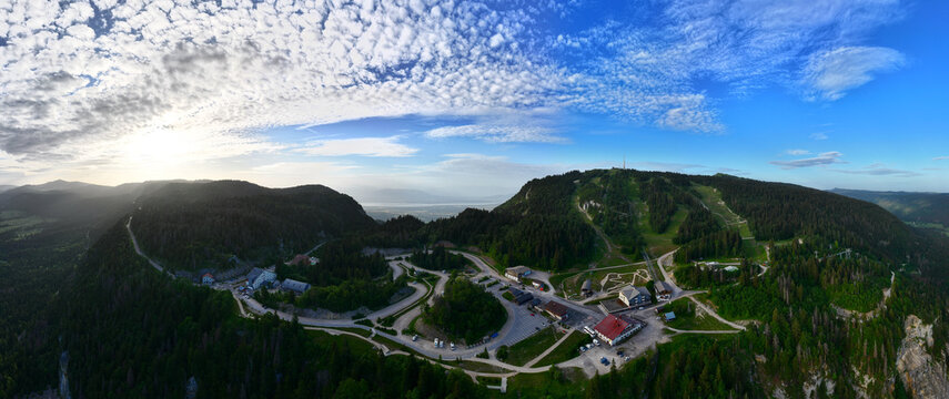 Aerial view of the Col de la Faucille mountain pass with winding roads, dense pine forests, and alpine resort buildings under a dramatic sunset sky in Gex, Auvergne-Rhone-Alpes, France.