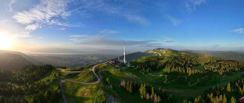 Aerial view of the Col de la Faucille mountain pass with a communication tower, winding roads, and dense forests overlooking a valley at sunset in Gex, Auvergne-Rhone-Alpes, France.
