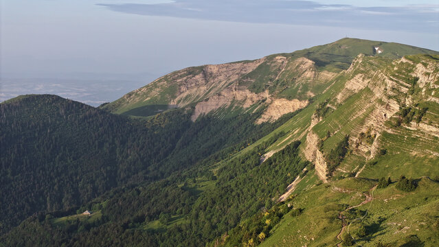 Aerial view of Col de la Faucille featuring rugged mountain ridges, lush green slopes, dense forests, and a winding trail in Gex, Auvergne-Rhone-Alpes, France.