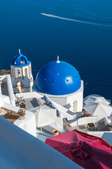 Iconic whitewashed Santorini architecture with blue domes overlooking calm Aegean waters and a passing boat. © Andrei Tomas