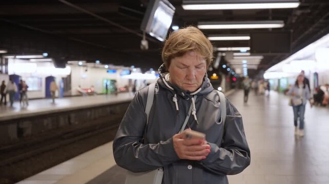 Senior woman traveler uses smartphone on an underground station platform while waiting for a commuter train arrival. Elderly passenger navigates public transit routes during a Paris trip.