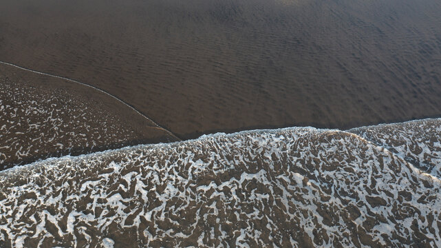 Aerial view of white sea foam washing over the dark volcanic black sand beach at Piha, Auckland, New Zealand.