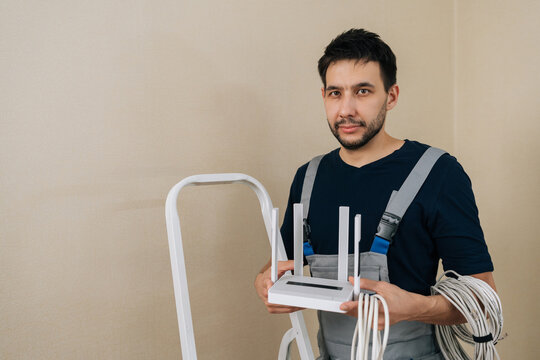 Professional male installer in uniform holding wireless router and cable for internet setup. He stands near ladder against neutral wall. Concept of connectivity and tech support.