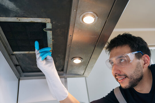Professional male worker with safety glasses and gloves performing inspection on dirty kitchen range hood filter, ensuring proper cleaning and maintenance of exhaust fan.
