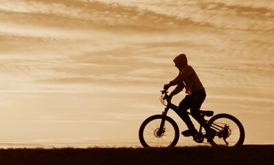 Fototapeta premium black silhouette of male young kid boy riding bicycle against yellow cloudy sky man on cycle in the nature at summer night Horizon hill line Unrecognizable person Summertime summer hot season