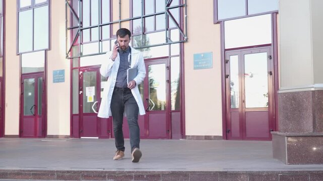 Young doctor in lab coat speak on mobile phone, leave hospital building stairs. Medical worker exiting hospital using cell phone. Medical university student walk outside building use smartphone.