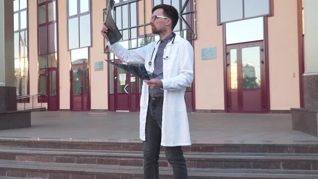 Doctor wearing lab coat stethoscope analyzing X-ray image outdoors stand near hospital main entrance. Young healthcare professional, internet radiologist exam mri x-ray lungs during COVID 19 epidemic.