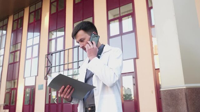 Male doctor in white lab coat talking on the phone with patient while examining his medical records outside the clinic. Physician consulting patient on test results. Personally delivering feedback.