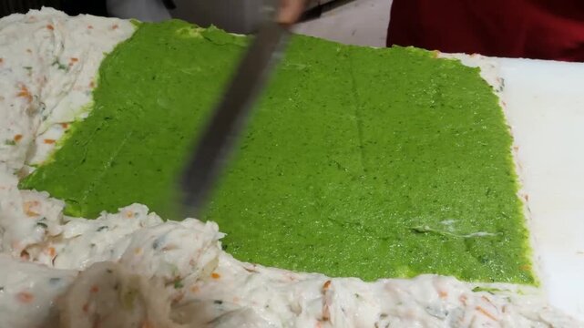 Close-up footage of a professional chef layering vibrant green vegetable-infused fish paste onto a white dough base to create traditional Korean fish cakes (Eomuk). This traditional street food proces