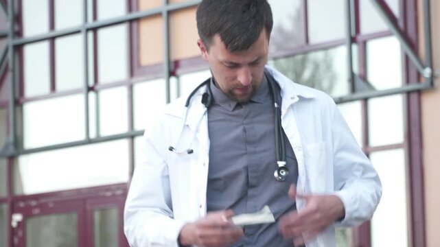 Topic is overtime work, heavy workload of medical workers. Young tired doctor went out to rest near entrance hospital after hard day at work, taking off his glasses, feels pain in eyes and drowsiness.