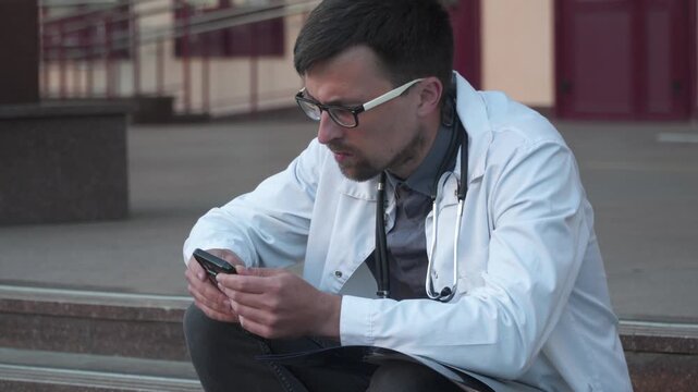 Worried tired doctor in white lab coat sit on floor outside hospital looking CT scan chest, corona virus, covid 19 concept. Frustrated medical professional, radiologist exam X-ray report pneumonia.