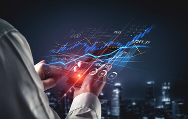 Obraz premium A close-up view of hands holding a smartphone displaying stock market data, with dynamic graphs and charts against a modern urban night skyline backdrop. Scalp
