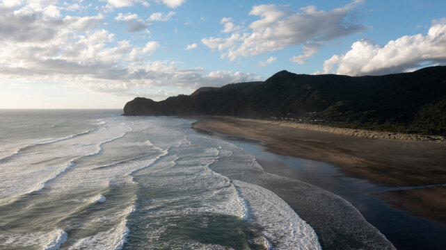 Aerial view of Piha Beach with dark volcanic sand, rolling ocean waves, and lush green hills under a cloudy sky in Piha, Auckland, New Zealand.
