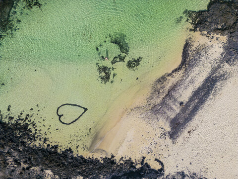 Aerial view of a heart shape drawn on the white sand beach surrounded by turquoise water and volcanic black rocks in El Cotillo, Canary Islands, Spain.