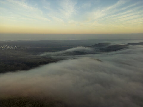 Aerial view of thick fog rolling over the volcanic hills and arid landscape of El Cotillo, Canary Islands, Spain.