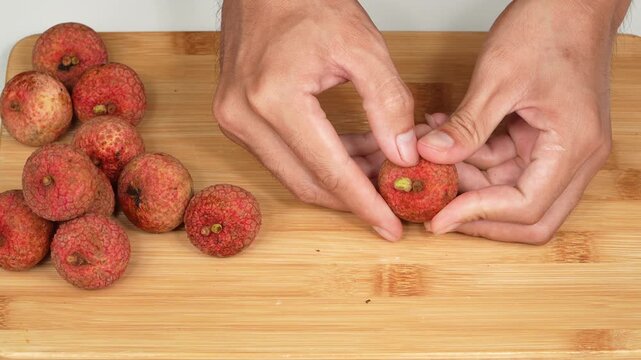 Macro shot of human hands peeling a fresh red lychee fruit on a wooden cutting board