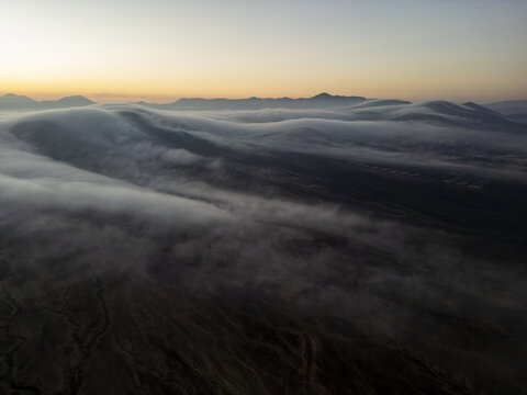 Aerial view of low-hanging clouds rolling over the volcanic hills during the golden hour in El Cotillo, Canary Islands, Spain.