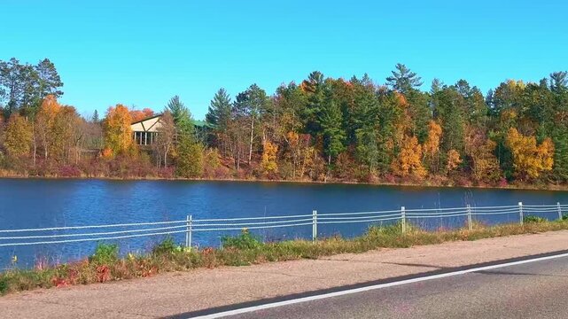 Driving POV alongside a Minnesota lake in peak autumn fall foliage near Walker, MN. Features vivid orange, gold, and red trees reflecting in blue water under a clear blue sky.
