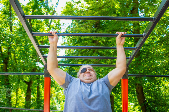 Middle-aged man with eyepatch doing pull-up on monkey bars in outdoor park fitness area