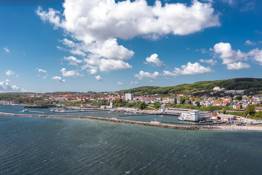 Elevated view of the Sassnitz waterfront in Mecklenburg-Vorpommern, Germany, showing harbor basin, promenade buildings, boats, and the Baltic Sea coastline in bright weather.