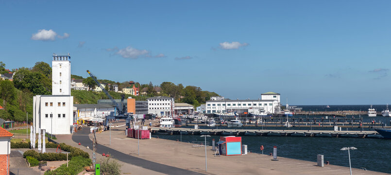 Panoramic waterfront scene in Sassnitz, Mecklenburg-Vorpommern, Germany, showing harbor promenade, seaside buildings, boats, and calm Baltic water on a sunny day.
