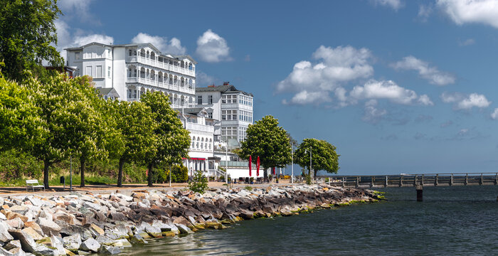 Waterfront view in Sassnitz, Mecklenburg-Vorpommern, Germany, with rocky shoreline, trees, white seaside buildings, and a pier under a blue summer sky.