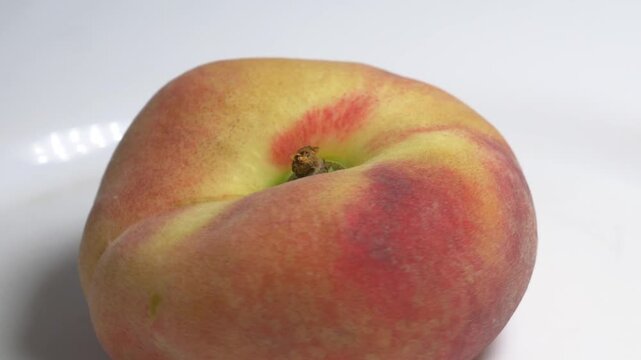 Close-up of a flat donut peach rotating on a white turntable against a light background, showcasing its unique shape and fuzzy skin under studio lighting.