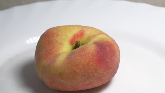 Close-up of a flat donut peach rotating on a white turntable against a light background, showcasing its unique shape and fuzzy skin under studio lighting.