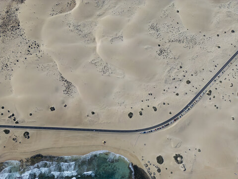 Aerial view of Parque Natural Dunas de Corralejo featuring vast sand dunes, a winding road with parked cars, and turquoise ocean waves in Corralejo, Canary Islands, Spain.