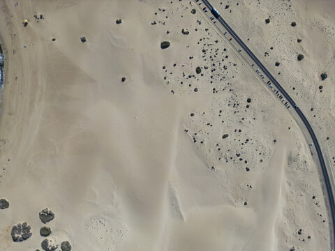 Aerial view of the Parque Natural Dunas de Corralejo with vast sand dunes, scattered desert vegetation, and a winding road with parked cars in Corralejo, Canary Islands, Spain.