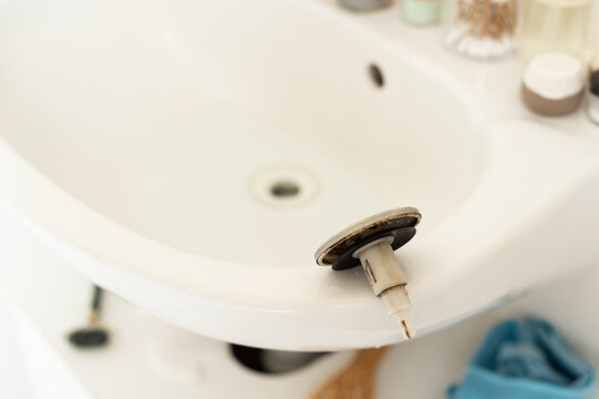 A silver and black metal sink strainer plug lies on the edge of a white ceramic basin. The bathroom drain in the background is being cleaned and cleared of a pipe clog.