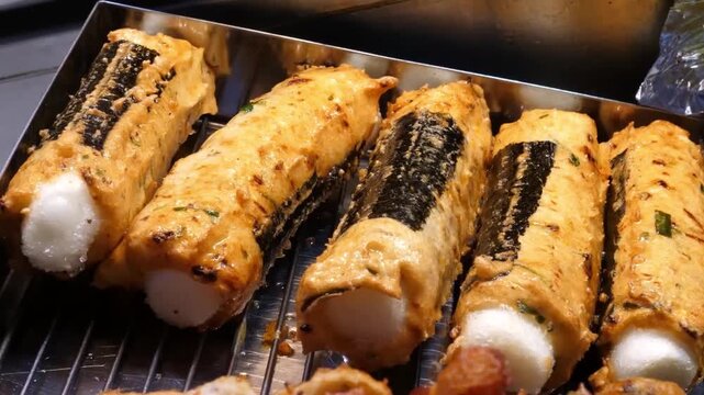 Close-up of freshly grilled Korean handmade fish cakes (eomuk) stuffed with rice cakes (tteok) and wrapped in seaweed. Various flavors including perilla leaf and spicy varieties are displayed on a met