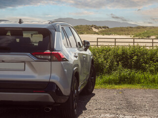 Fototapeta premium Silver SUV parked on gravel near fence. Mountainous landscape stretches in background. Green bushes line the parking lot. Sky shows scattered clouds. Outdoor travel and tourism. Rented vehicle.