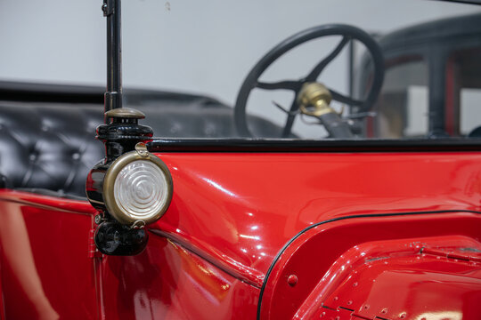 A detailed view of a historic automobile side lamp with the steering wheel visible in the background. The image showcases vintage vehicle design and craftsmanship.