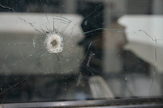 Close up of a cracked vintage car window with a visible bullet hole impact. The damaged glass shows radial fracture patterns and signs of historical wear.