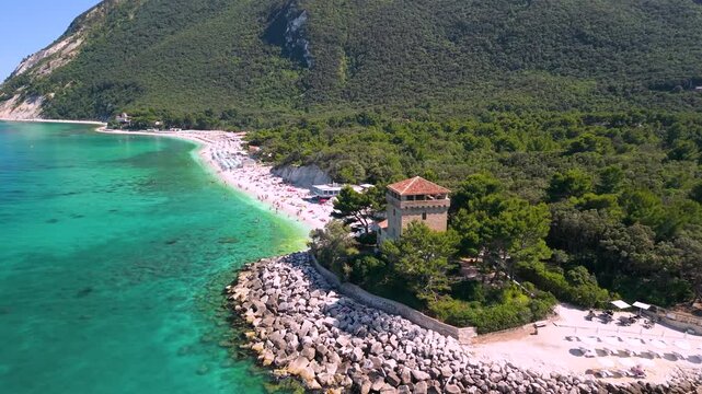 Portonovo Marche Italy 4k drone of Torre Clementina beach. Aerial tracking flight over white pebbles coast to historic Santa Maria church with canoes in tropical turquoise sea water b-roll