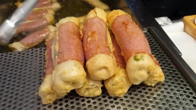 A close-up shot of Korean street food fish cakes wrapped in ham and freshly fried on a metal rack.