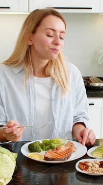 Middle-aged woman eating trout fish with avocado nuts and vegetables for keto diet in kitchen at home feeling satisfied. Young girl choosing low carb meal for healthy fit nutrition and weight loss