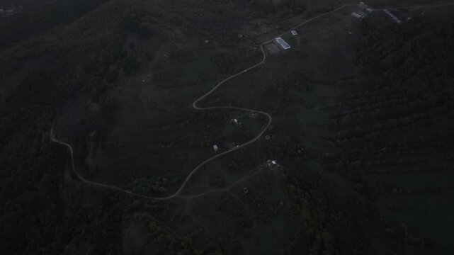 A dark high angle aerial view of a winding gravel road traversing a mountain forest valley with scattered houses during a gloomy twilight or dawn in the countryside.

